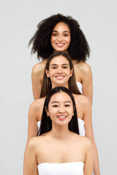 Grupa trzech różnych pięknych kobiet uśmiechających się do kamery, stojących i pozujących na szarym tle studia. Pionowy portret urody młodych dam - Zdjęcie, obraz