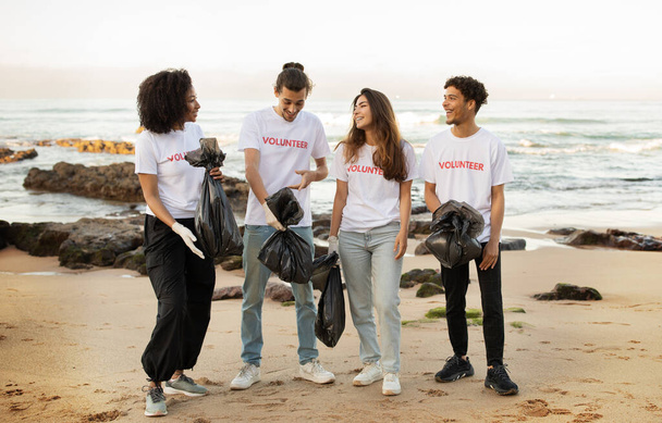Χαρούμενοι νέοι και διαφορετικοί άνθρωποι εθελοντές σε μπλουζάκια με σακούλες σκουπιδιών καθαρίζουν τα σκουπίδια στην παραλία της θάλασσας, διασκεδάζουν σε εξωτερικούς χώρους, σε όλο το μήκος. Οικολογική διατήρηση, περιβάλλον, προστασία του πλανήτη - Φωτογραφία, εικόνα