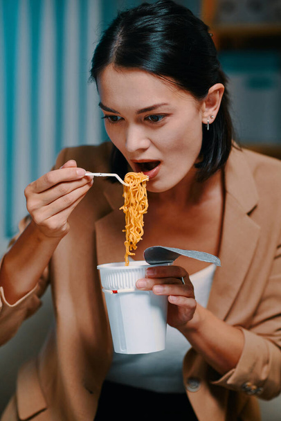 Businesswoman eating instant ramen when reading document on computer screen in dark office - Photo, Image