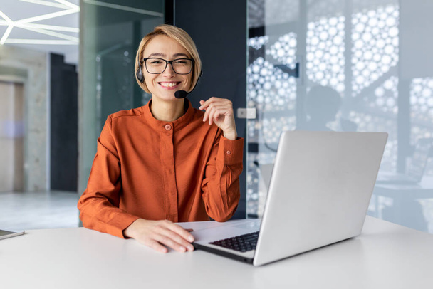 Portrait of successful online customer support office worker, satisfied business woman at workplace with video call headset and laptop smiling and looking at camera. - Photo, image
