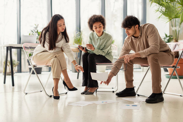 Power Of Teamwork. Three Multiethnic Colleagues Brainstorming And Discussing Business Project Looking At Papers On Floor Sitting In Chairs In Modern Office Indoors. Corporate Meeting Concept - Foto, Imagem