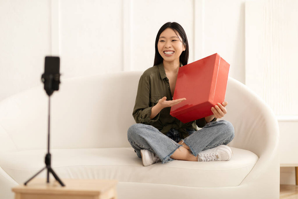 Onnellinen iloinen kaunis nuori aasialainen nainen vaikuttaja tai kuuluisa bloggaaja, jolla on punainen cardbox-paketti, istuu sohvalla kotona, streaming samalla kun tarkistetaan hänen upouudet kengät tai kangas, purkaminen toimitus - Valokuva, kuva