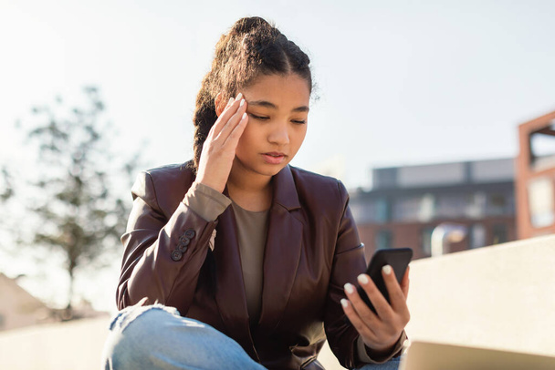 Worried mixed race lady reading message on smartphone while sitting outdoors, confused female student looking at cellphone screen and frowning, received unpleasant sms - Photo, image