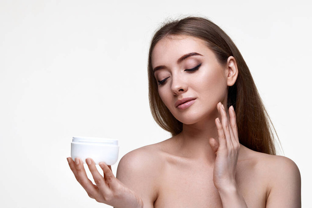 beautiful smiling girl applying skin care cream on her face and holding jar of cosmetic product on white background - Foto, imagen