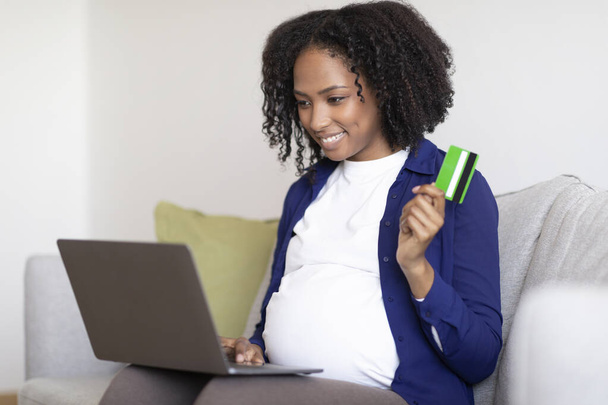 Alegre señora embarazada afroamericana milenaria con gran vientre con computadora portátil muestra tarjeta de crédito en el sofá en el interior de la sala de estar. Esperando bebé, compras en línea, gran venta y reembolso - Foto, imagen