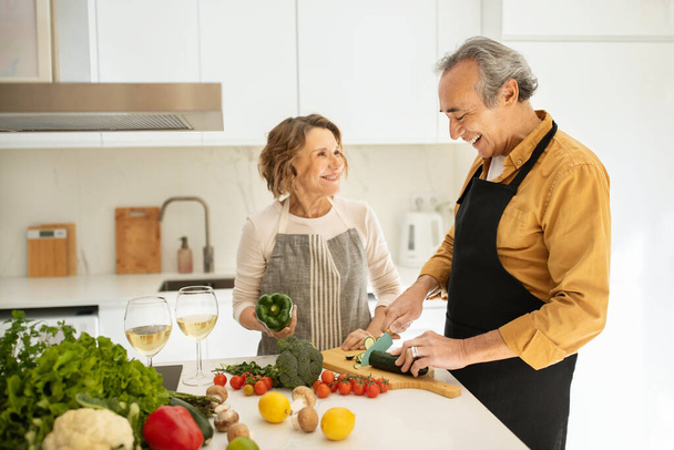 Glückliche ältere Eheleute kochen zusammen und reden, Frau und Mann bereiten Gemüsesalat zu, kochen gemeinsam Abendessen, Kücheneinrichtung, Kopierraum - Foto, Bild