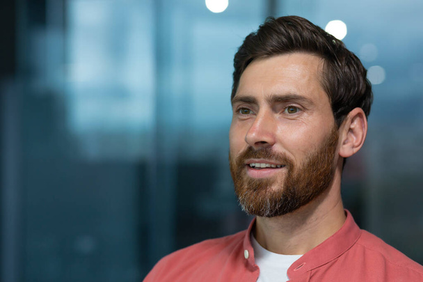 Close-up photo. Portrait of a successful and smiling young man with a beard and wearing a red shirt sitting in the office and confidently looking away. Thinks, dreams, poses. - Valokuva, kuva