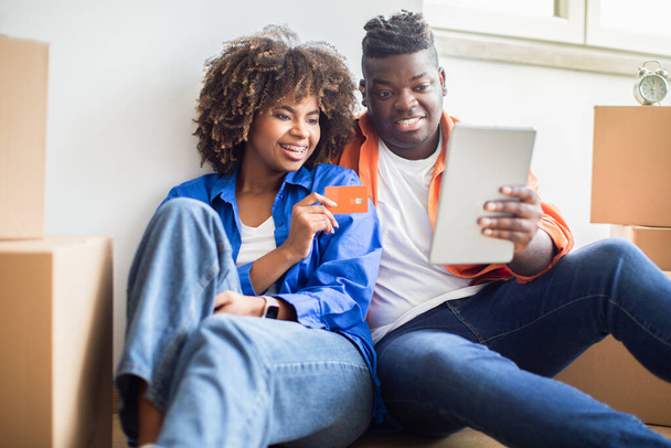 Pareja familiar negra con tableta digital y tarjeta de crédito que elige muebles en línea después de mudarse a un nuevo apartamento, sonriendo a los cónyuges afroamericanos con la pestaña Compras por computadora en Internet, Primer plano - Foto, imagen