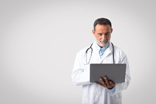 Serious confident european senior man doctor in white coat with stethoscope typing on laptop on gray background, free space. Professional work, medical care remotely, exam, ad and offer - Fotografie, Obrázek