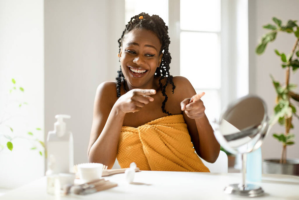 Power of daily skincare. Happy african american lady pointing fingers at her reflection in small mirror, sitting at dressing table in bedroom while making daily beauty routine - Photo, Image