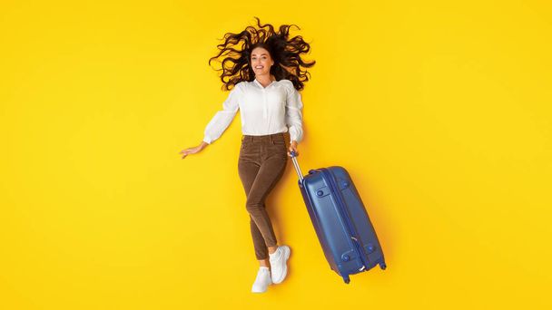Reisaanbieding. Vrolijke jonge dame poseren met blauwe koffer reclame reisbureau liggend op gele achtergrond, bovenaanzicht. Volledige shot van toeristische vrouw met bagage. Vakantie concept. Panorama - Foto, afbeelding