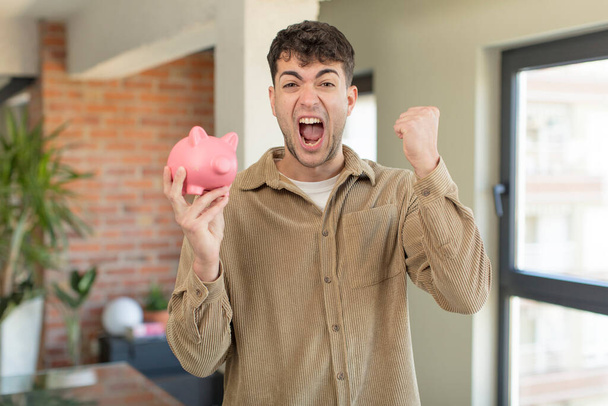 young handsome man feeling shocked,laughing and celebrating success. piggy bank concept - Fotoğraf, Görsel