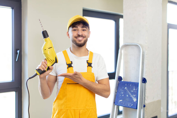 young handsome man smiling cheerfully, feeling happy and showing a concept. handyman and drill concept - Foto, Imagen