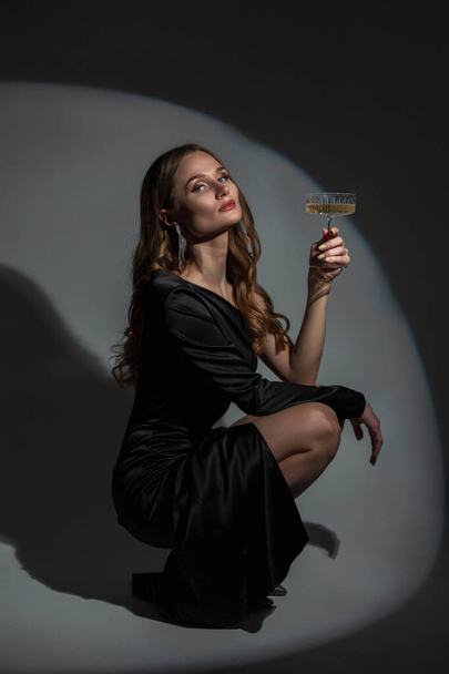 Mooie stijlvolle elegante vrouw met een diamanten oorbel in een mode zwarte avondjurk met een glas champagne zit en poseert in de studio op een donkere achtergrond met hard licht. Lady op het feest - Foto, afbeelding
