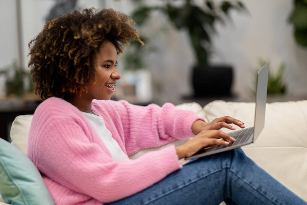 Cheerful attractive curly young black woman chilling at home, sitting on couch with legs up, using modern pc laptop, websurfing, reading online blog, gaming, side view, copy space - Photo, image