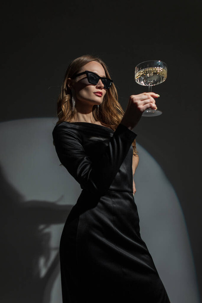 Preciosa mujer chic joven de moda con gafas de sol frescas en un elegante vestido elegante negro con una copa vintage de champán sobre un fondo oscuro en una fiesta. Bella dama celebra un evento - Foto, imagen
