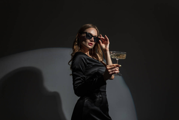 Модна красива елегантна модна жінка носить прохолодні сонцезахисні окуляри в модній вечірній чорній сукні з келихом шампанського на вечірці. Шикарна красива дама святкує подію
 - Фото, зображення