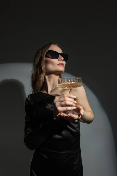 Hot elegant pretty stylish woman with sunglasses in black fashion dress holds vintage glass of champagne at party. Chic beauty lady celebrate - Фото, зображення
