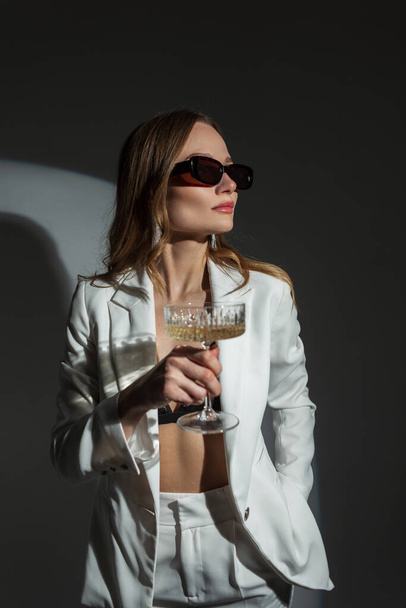 Coole Mode hübsche schicke Frau mit Vintage-Sonnenbrille in einem modischen weißen Anzug mit einem Glas Champagner auf dunklem Hintergrund auf einer Party - Foto, Bild