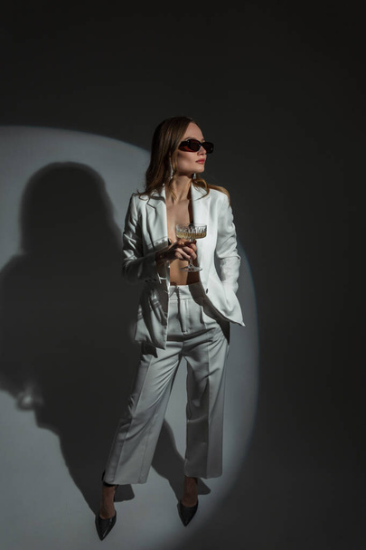 Elegant beautiful lady with sunglasses in a fashion white suit with a vintage glass of champagne at a party - Fotó, kép