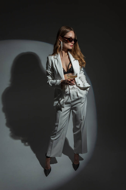 Fashionable beautiful woman in a stylish elegant white suit with a blazer and a bra with heels holds a glass of champagne and poses in the studio on a dark background with circular light. Pretty lady - Fotografie, Obrázek