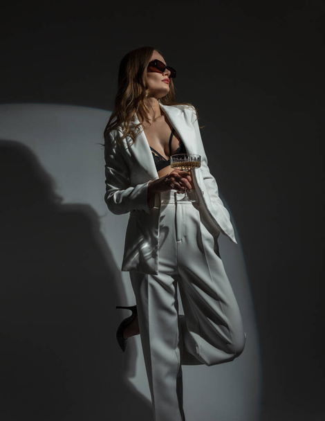 Fashion beautiful chic woman with cool sunglasses in a stylish white suit with a blazer and a bra with a glass of champagne on a dark background with a round light at a party. Lady celebrating - Fotografie, Obrázek
