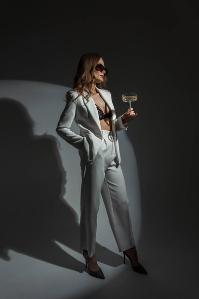 Piękna elegancka młoda elegancka kobieta w okularach przeciwsłonecznych w białym modnym garniturze z marynarką, top i obcasami ze szklanką szampana na ciemnym tle w studio. Pani świętuje wydarzenie. - Zdjęcie, obraz