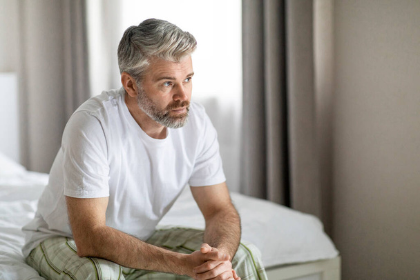 Pensativo pensativo infeliz guapo de pelo gris barbudo maduro caucásico hombre con pijama sentado en la cama en casa, mirando el espacio de copia, sintiéndose solo, pasando por el divorcio - Foto, Imagen