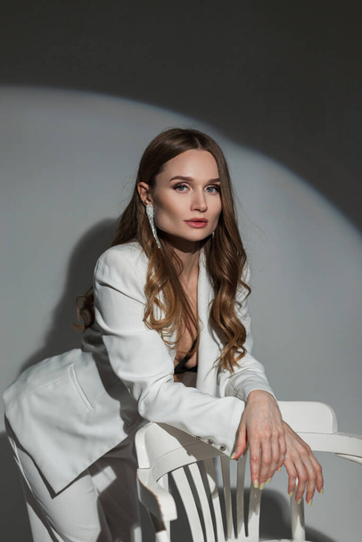 Schöne modische Dame im weißen eleganten Anzug steht neben einem weißen Stuhl auf dunklem Hintergrund im Studio bei einer Party - Foto, Bild