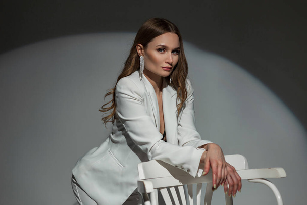 Stylish elegant business lady with a diamond earring in a fashionable white suit with a bra posing near a white chair on a dark background in the studio. Pretty chic woman at party - 写真・画像