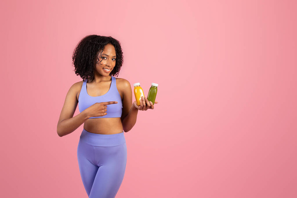 Milenyum siyahi kadın spor kıyafetleri içinde pembe arka planda, stüdyoda meyve suyu ya da meyve suyu dolu kavanozlar tutuyor. Spor, diyet ve sağlık içeceği ve vücut bakımı için tavsiye - Fotoğraf, Görsel