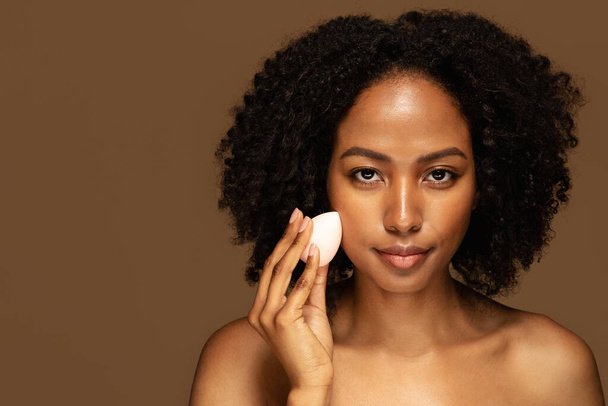 Young pretty african american woman taking off makeup with cotton wipe sponge. Smiling lady cleaning face isolated over background. Black young woman cleansing face, daily healthy beauty routine - Foto, Imagem