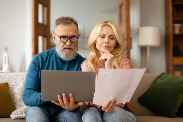 Balancing the Budget. Serious Mature Spouses Thoughtfully Reviewing Paper Bills And Using Laptop, Managing Finances Online Sitting On Sofa At Home. Household Paperwork And Retirement Concept - Foto, Imagen