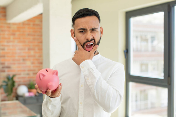 young  adult man with mouth and eyes wide open and hand on chin. piggybank concept - Foto, afbeelding