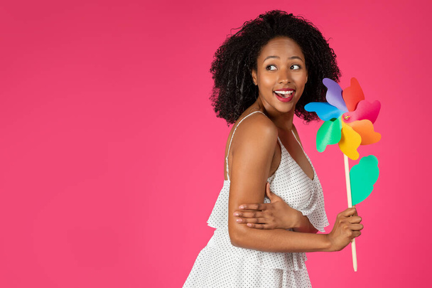 Vrolijke schattige jonge Afrikaanse Amerikaanse krullende vrouw met windmolen bloem genieten van vrije tijd, vakantie, weekend, kijken naar vrije ruimte, geïsoleerd op roze studio achtergrond. Verkoop, plezier, vrijheid en mode - Foto, afbeelding