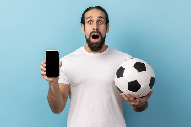 Portrait of amazed man with beard wearing white T-shirt holding soccer ball and smartphone empty black display, ticket booking for championship. Indoor studio shot isolated on blue background. - Photo, image