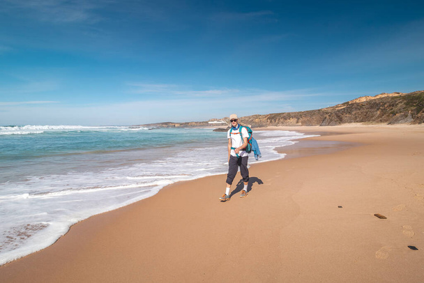Backpacker loopt langs Praia do Almograve met een glimlach op zijn gezicht. De vreugde van het verhuizen en het ontdekken van nieuwe plaatsen. Odemira, West-Portugal. Wandelen langs de Vissersroute, Rota Vicentina. - Foto, afbeelding