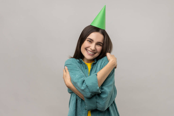 I love myself. Woman with dark hair and funny party cone on head, congratulating and embracing herself on birthday, wearing casual style jacket. Indoor studio shot isolated on gray background. - Foto, imagen