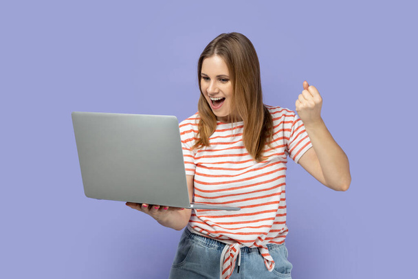 Portrait of extremely happy beautiful blond woman wearing striped T-shirt working on laptop, looking at screen and clenched fist, showing yes gesture. Indoor studio shot isolated on purple background. - Foto, Imagem