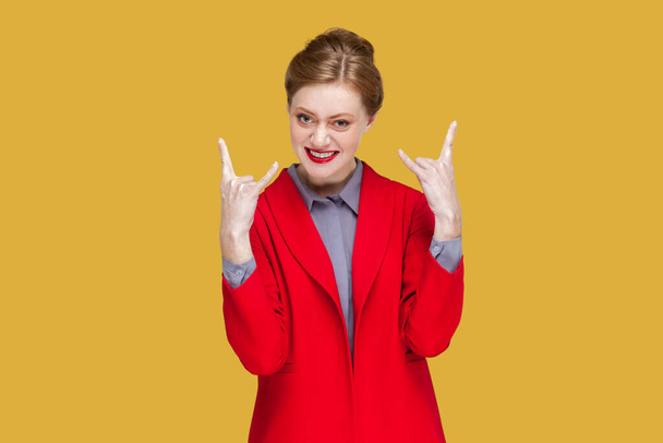 Portrait of excited crazy woman with red lips standing showing rock and roll gesture, having fun on festival, wearing red jacket. Indoor studio shot isolated on yellow background. - Fotografie, Obrázek