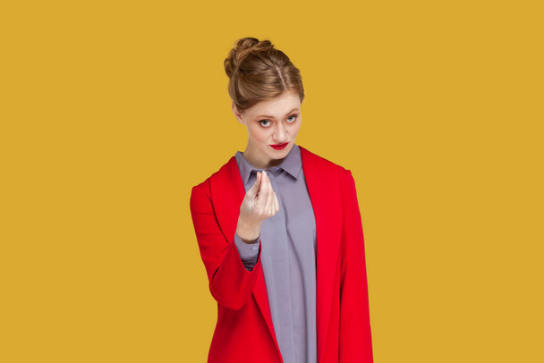 Portrait of serious young adult woman with red lips standing and showing money gesture, asking to give her salary, wearing red jacket. Indoor studio shot isolated on yellow background. - Фото, изображение