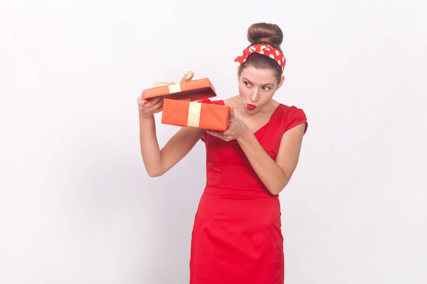 Portrait of curious attractive woman wearing red dress and head band holding present box, looking inside with interest, having gift on her birthday. Indoor studio shot isolated on gray background. - Photo, Image