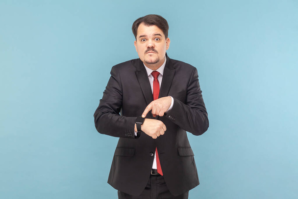 Portrait of serious man with mustache standing looking at camera, pointing at his smartwatch, deadline, wearing black suit with red tie. Indoor studio shot isolated on light blue background. - Фото, изображение