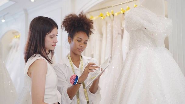 Twee professionele vrouwen mode-ontwerper met behulp van tablet voor ontwerp witte trouwjurk in de trouwstudio - Foto, afbeelding