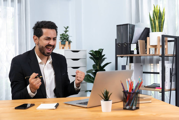 Excited and happy businessman dressed in black formal suit, raise his arm in celebratory gesture at his office desk, after successful job promotion, energy and joy as business winner. Fervent - Fotoğraf, Görsel