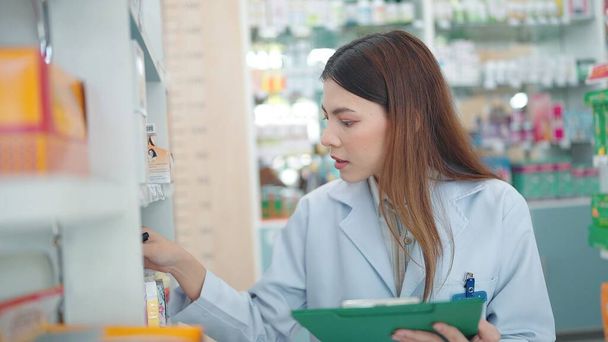 Profesional mujer asiática farmacéutico vuelve a comprobar los productos en la farmacia y el uso de portátil para escribir información - Foto, Imagen