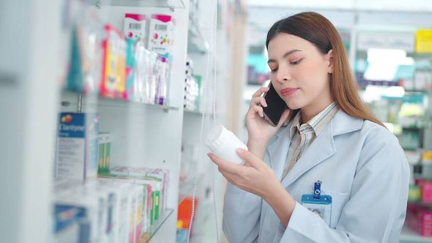 Profesional mujer asiática farmacéutica ayudando al cliente con la recomendación de la medicina hablando con el cliente en el teléfono inteligente en la farmacia - Foto, Imagen