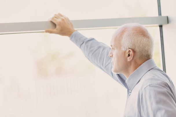 A gray-haired, almost balding man looks out of a large, bright, modern window. He has his hand resting high on the frame and is looking at something in the distance, perhaps thoughtful or worried. - Photo, image