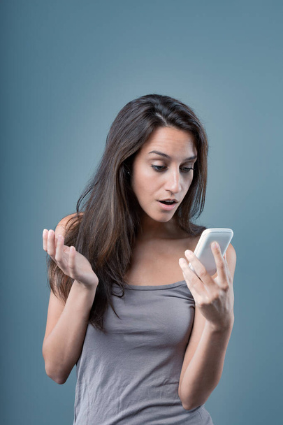 Emotional portrait of a young woman isolated on a neutral background. Surprised on the phone. Girl in a shocking moment due to something on her cellphone, with a bewildered hand gesture. Long brown ha - Фото, изображение