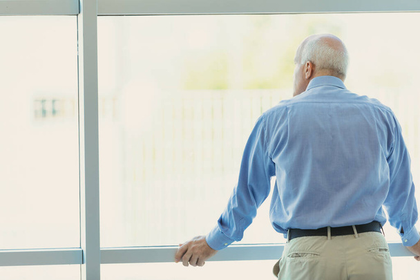 Un homme aux cheveux gris, presque chauve, regarde par une grande fenêtre lumineuse et moderne, nous tournant le dos. Il a les mains basses sur le cadre et regarde quelque chose au loin, peut-être th - Photo, image